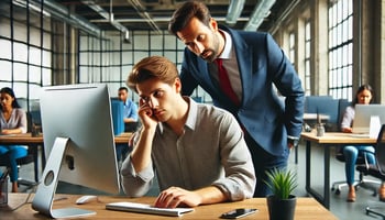 A professional office setting where a manager closely oversees an employee's work on a computer screen. The employee appears frustrated or overwhelmed, illustrating the concept of micromanagement in a modern workplace.